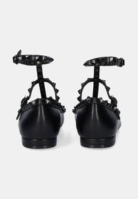 Zapatos planos de cuero negro con punta afilada, acentos de tachuelas alrededor del tacón y una correa con hebilla en el tobillo. Textura suave, diseño minimalista.