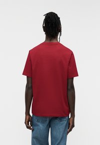 Κόκκινο βαμβακερό μπλουζάκι με κοντά μανίκια, στρογγυλή λαιμόκοψη και απλό σχέδιο. Φορεμένο με ανοιχτόχρωμο μπλε τζιν, από την πίσω πλευρά.