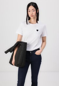 Camiseta blanca de algodón con un pequeño gráfico de corazón negro. Combinada con jeans azul oscuro y una chaqueta negra acolchada sostenida con una mano.