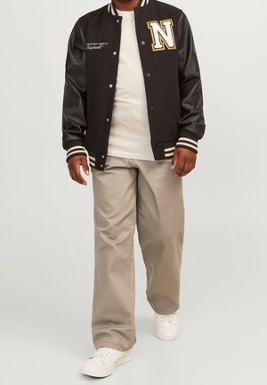 Mand iført sort varsityjakke med bogstavet N, hvid t-shirt, beige bukser og hvide sneakers, gående mod en ensfarvet baggrund.