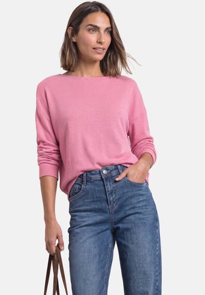 Femme portant un pull rose et un jean bleu, tenant un sac à bandoulière marron, une main dans la poche, debout devant un fond blanc.