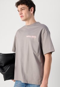 Pilkas medvilnės T-shirt su laisva iškirpte ir trumpomis rankovėmis, su rožinės spalvos spauda ant krūtinės. Modelis laiko juodą rankinę.