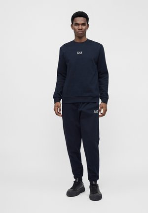 Homme debout, portant un sweat-shirt bleu marine et un pantalon de survêtement assorti avec un logo blanc EA7, et des baskets noires sur un fond clair uni.