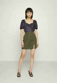 Blusa escura com padrão floral e mangas bufantes, combinada com uma saia mini verde com uma fenda lateral. Modelo usa sandálias pretas de tiras.