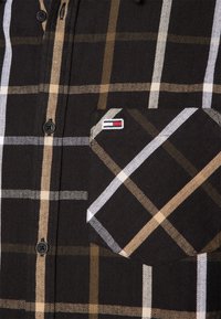 La camisa de cuadros negra presenta cuadros en marrón, blanco y beige, cierre de botones y un bolsillo con parche que incluye un pequeño detalle del logo.