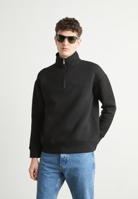 Hanorac negru cu fermoar si guler înalt, având un logo subtil Calvin Klein. Asortat cu blugi de un albastru deschis si ochelari de soare negri.
