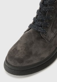 Stivaletto alla caviglia in suede grigio scuro con superficie texturizzata, suola piatta in gomma nera e lacci in lana nera attraverso occhielli metallici.