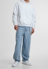 Felpa bianca con cappuccio e tasca a marsupio, abbinata a jeans a gamba larga di un leggero blu con accessorio a catena e sneakers bianche.