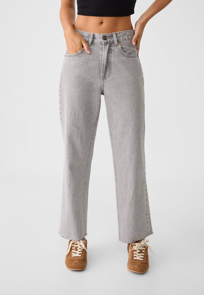 Jean à taille haute à coupe droite en denim gris clair. Ourlet effiloché, design à cinq poches et détails de couture minimalistes. Porté avec des baskets beige.