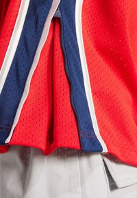 Maillot de sport en maille rouge et marine avec des bordures blanches, présentant une texture perforée et une fermeture zippée partielle. Short gris uni partiellement visible.