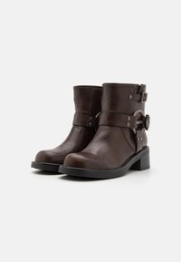 Botine din piele maro, cu vârful rotund, curea cu cataramă și finisaj texturat. Prezintă un toc cub, mic și accente metalice.