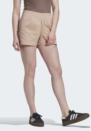Personne portant un short beige à taille élastique, un haut marron sans manches et des baskets noires avec des bandes blanches, debout sur un fond clair.