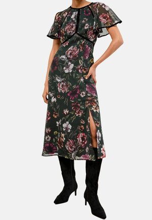 Kvinde iført en mørk blomster midi-kjole med gennemsigtige korte flagrende ærmer og en sideslid, kombineret med sorte knæhøje støvler med hæl.