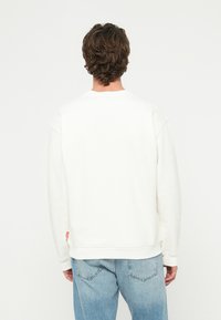 Muž s krátkymi kučeravými vlasmi, oblečený v jednoduchom bielom pulóvri a svetlomodrých džínsach, stojaci chrbtom k bielej jednoliatej stene.