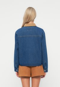 Denim jacket featuring a blue wash with a tan corduroy collar, long sleeves, and button closure. Seen from the back, paired with brown shorts.