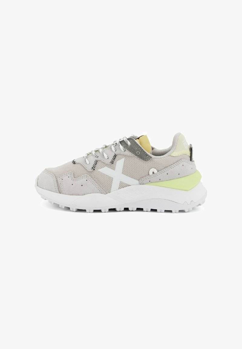 Zapatilla deportiva gris y blanca con materiales de malla y ante, un acento verde en el talón, y una suela blanca con patrón. Cuenta con cordones reflectantes.