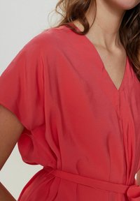 Rochie coral cu decolteu în V, mâneci scurte și un brâu legat la talie. Fabricată dintr-un material ușor, cu o textură netedă.
