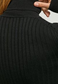 Svart ribbad polotröja med vertikala linjer, hög krage och en mjuk textur, med närbild på halsringningen.