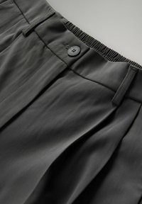Dunkelgraue Hose mit elastischem Bund, Knopfverschluss, Gürtelschlaufen und Faltendetail auf der Vorderseite vor einem hellgrauen Hintergrund.
