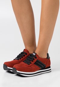Rote Wildleder-Sneaker mit schwarzen Akzenten, weißen Streifen an der Sohle und schwarzen Schnürsenkeln. Verfügt über ein strukturiertes Design und ein Logo-Detail an der Seite.