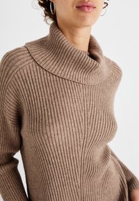 Gros plan sur une personne portant un pull en maille côtelée beige avec un col large roulé et des boucles d'oreilles en argent.