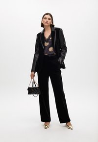 Traje de terciopelo negro con solapas de satén, superpuesto a una blusa oscura con puntos dorados y bordes de encaje. Los accesorios incluyen un pequeño bolso negro acolchado.