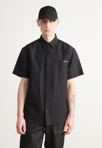 Camisa negra de manga corta abotonada en mezcla de algodón con bolsillo y logotipo bordado. Combinada con pantalones de mezclilla oscuros y una gorra negra.