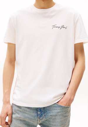 Biała bawełniana koszulka z krótkim rękawem, okrągłym dekoltem i czarnym logo w formie napisu na lewej piersi. W zestawieniu z jasnoniebieskimi dżinsami.