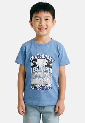 Jeune garçon souriant portant un T-shirt Lee Cooper à thème skateboard bleu et un jean bleu clair sur un fond uni.