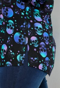 Schwarzes Shirt mit einem Muster aus bunten Schädeln und Sternen. Die Schädel sind in Blau- und Lilatönen auf schwarzem Hintergrund gehalten.