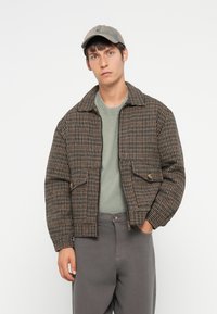 Brown checkered jacket with a collar, two front pockets, and ribbed cuffs. Worn over a green shirt and gray trousers, paired with a cap.