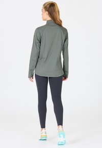 Lichte, olijfgroene sportjack met hoge kraag, lange mouwen en een aansluitend ontwerp, gecombineerd met marineblauwe leggings en sportschoenen.