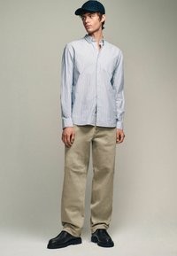 Light blue and white striped button-up shirt, tan pants, black shoes, and a dark cap. Casual fit with a smooth fabric texture.
