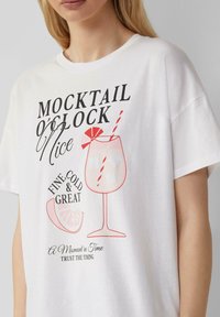 T-shirt blanc à manches courtes avec un design graphique représentant un verre à cocktail, du texte dans différentes polices et couleurs, ainsi qu'un détail de quartier de lime et de paille.