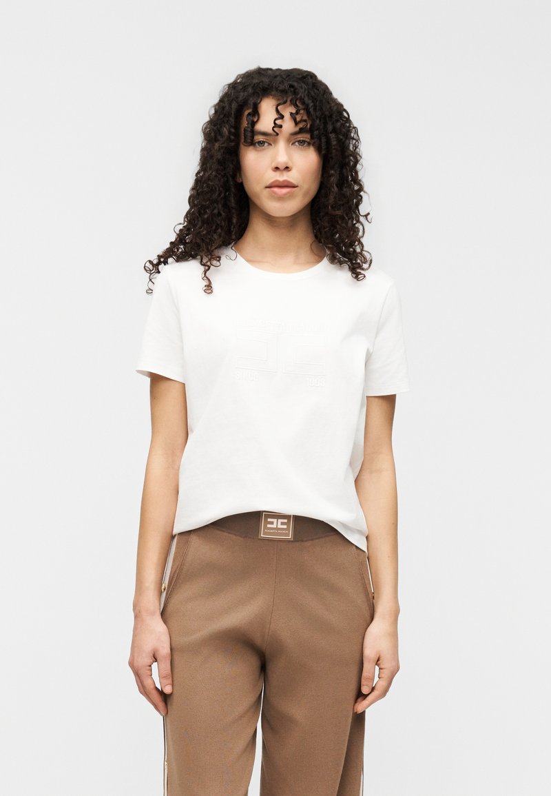 Λευκό βαμβακερό t-shirt με κοντά μανίκια και ανάγλυφο λογότυπο στο μπροστινό μέρος, σε συνδυασμό με καφέ παντελόνι με ανοιχτό καφέ ζώνη μέσης και λογότυπο.