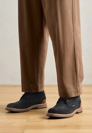 Black suede ankle boots with a contrasting brown heel, round toe, and tan sole. Paired with loose, brown trousers.