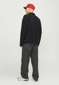 T-shirt noir à rayures, manches longues, et pantalon baggy gris foncé. Le mannequin porte une casquette rouge et des baskets claires avec des semelles texturées.