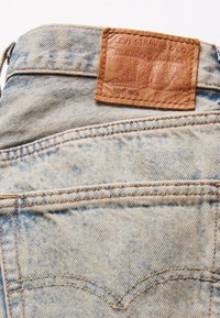 Vaqueros de mezclilla azul claro con un parche de cuero marrón texturizado que presenta detalles de la marca en relieve, y costuras en contraste en los bolsillos traseros.