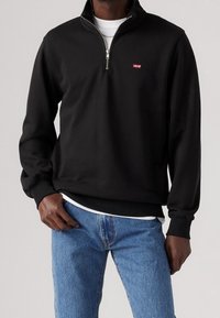 Uomo che indossa un pullover nero Levi's a zip quarter sopra una maglietta bianca con jeans azzurri, in posa con una mano in tasca, sfondo neutro.