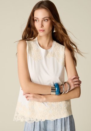 Jeune femme aux longs cheveux portant un top en dentelle blanche sans manches et une jupe bleu-gris, accessoirisée de larges bracelets bleus et argentés, regardant sur le côté.