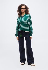 Γυναίκα που φοράει μακρυμάνικο πόλο με πράσινες και λευκές ρίγες, σκούρα τζιν με φαρδιά πόδια και μπρελόκ, λευκά παπούτσια και σκούρα γυαλιά ηλίου, στέκεται σε απλό φόντο.