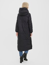 Manteau de courtepointe noir, long, avec capuche, présentant un design matelassé et des coutures latérales. Accompagné d'un jeans bleu et de bottes à talons noires.