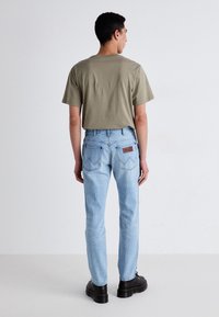 Jeans de color azul claro con bolsillos traseros que presentan un patrón de costura en forma de chevron, combinados con una camiseta de manga corta en color caqui y zapatos de plataforma negros.
