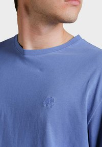 Světle modré bavlněné tričko s kulatým výstřihem, na levé straně hrudi má malé vyšívané logo slona. Měkká textura, volný střih.