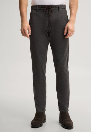 Uomo che indossa pantaloni slim fit grigio scuro con coulisse e stivali in pelle scamosciata marrone scuro, in piedi su uno sfondo neutro.