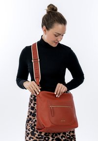 Bruine crossbodytas van getextureerd leer met een voorvak met rits en een verstelbare schouderband met patroon. Het ontwerp is rechthoekig met afgeronde hoeken.