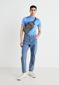 Gray crossbody bag with front pocket, worn over a blue athletic shirt and paired with light blue jeans and canvas sneakers. Smooth textures.