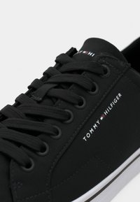 Baskets noires avec lacets noirs, semelle blanche, et logo ainsi que détail du drapeau "Tommy Hilfiger" sur le côté et la languette.