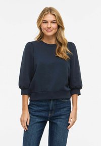 Femme blonde aux cheveux ondulés lâchés, portant un chemisier bleu marine à manches bouffantes et un jean bleu, debout devant un fond blanc uni.