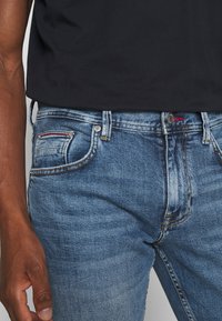 Vaqueros de mezclilla azul con textura desgastada, que presentan un bolsillo delantero con detalles de costuras en rojo y blanco y un botón plateado en la cintura.